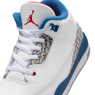 Kids white sneaker with laces, blue Jumpman emblem, red eyelets, and cement grey print.