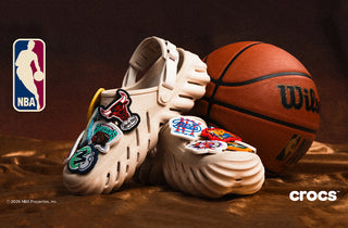 White Crocs shoes with colorful patches next to a basketball on a dark background, featuring NBA logo.